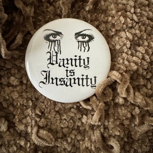 May include: White button with a black and white image of two eyes with tears and the text "Vanity is Insanity".