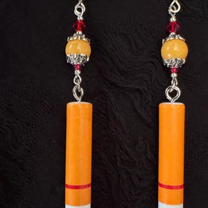 Op de afbeelding: Een paar novelty oorbellen die op sigaretten lijken. Elke oorbel heeft een zilveren haak, rode kralen, een gele kraal en een cilindrische oranje en witte hanger met een rode band. De uiteinden zijn donkerrood.