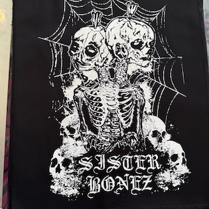 May include: Black patch featuring a white graphic of two conjoined skulls, a skeleton, and a spiderweb design. The text "SISTER BONEZ" is printed in a gothic font. The patch is approximately 8 inches wide.