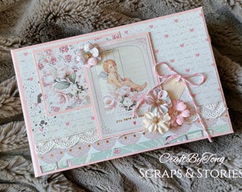 May include: A rectangular handmade card featuring a central illustration of a cherub surrounded by pink roses, accented with layered paper flowers, white lace trim, pink twine, and the printed text you have my heart on a light pink background.