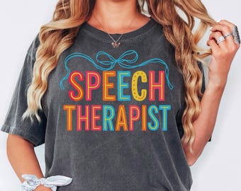 May include: Dark gray t-shirt with the words "SPEECH THERAPIST" in colorful, outlined letters. The text is in a bold, playful font, with a blue bow above. The word "pepper" is in the bottom right corner.