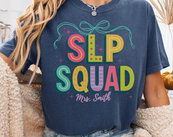 May include: A navy blue t-shirt with the words "SLP SQUAD" in colorful, bubble letters. The shirt also features the name "Mrs. Smith" and the word "Denim" at the bottom. A bow and stars decorate the top of the text.