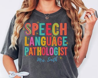 May include: Dark gray t-shirt with the words "SPEECH LANGUAGE PATHOLOGIST" in colorful, retro-style lettering. The name "Mrs. Smith" is below the text. The word "pepper" is in the bottom right corner.