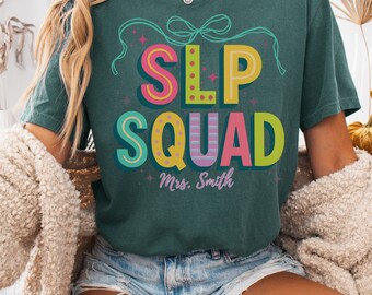 May include: A teal-colored t-shirt with the words "SLP SQUAD" in colorful, retro-style lettering. Below the text is "Mrs. Smith" in a smaller font. The shirt also has a bow graphic and the words "Blue Spruce" at the bottom.