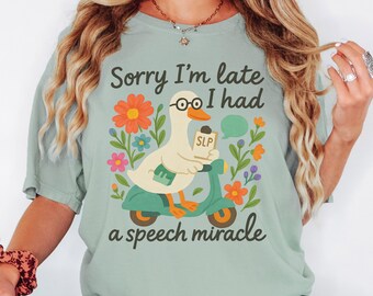 May include: A light green t-shirt with the text "Sorry I'm late I had a speech miracle." The graphic features a duck wearing glasses and riding a teal scooter. Colorful flowers and a speech bubble complete the design.