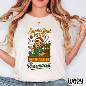 May include: Ivory-colored t-shirt featuring a cartoon pharmacist in a Santa hat, with the text "Santa's Real MVP" and "Pharmasleigh The Pharmacist." The design includes gift boxes, medicine bottles, and a shelf of products.