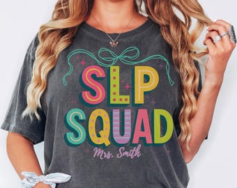 May include: Dark gray t-shirt with the words "SLP SQUAD" in colorful, block letters. The shirt also features the name "Mrs. Smith" and a bow design above the text. The word "pepper" is written in the bottom right corner.