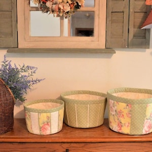 May include: Three fabric storage baskets with floral and polka dot patterns. The baskets are lined with a cream-colored fabric and have green trim. The baskets are arranged in a row on a wooden surface.