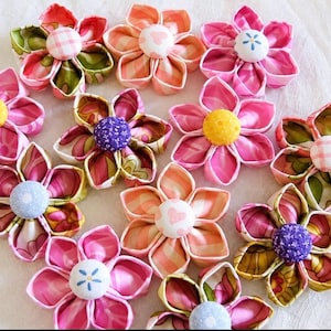May include: A collection of eleven fabric flowers, each featuring five petals made from various patterned textiles in shades of pink, green, orange, and yellow. Each flower has a central fabric-covered button with diverse prints.