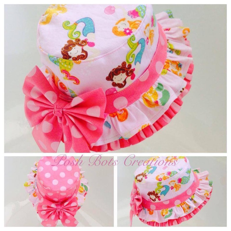 May include: Pink sun hat with a white polka dot bow and a pink ruffle trim. The hat is decorated with a mermaid print featuring colorful mermaids and seashells.