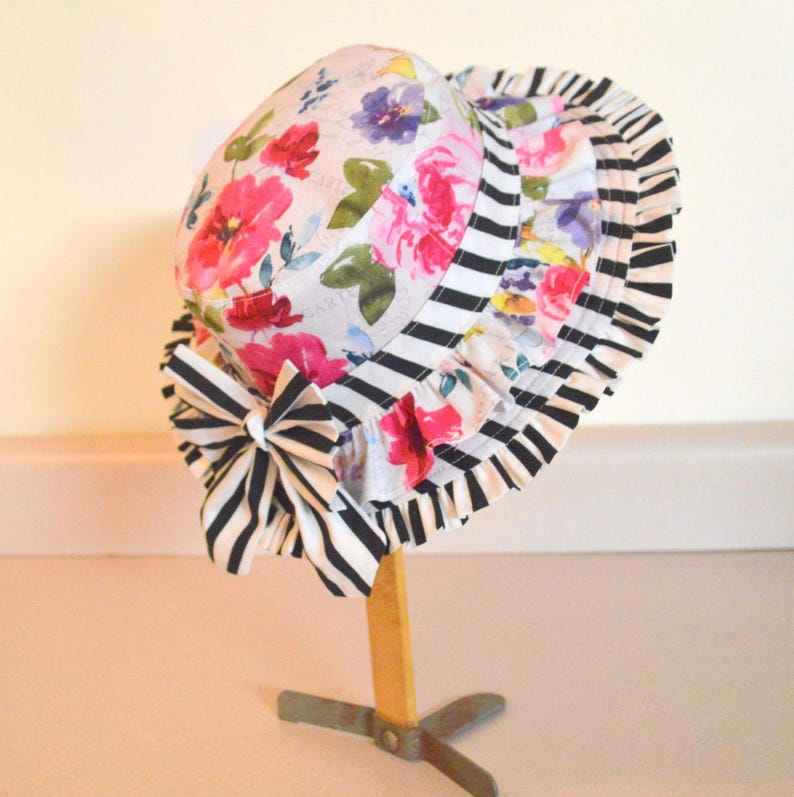 May include: A white sun hat with a black and white striped trim and a floral print. The hat has a ruffle around the brim and a bow at the back.