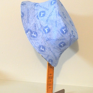 May include: A blue bucket hat with a white John Deere logo pattern. The hat is on a wooden stand with a ruler.