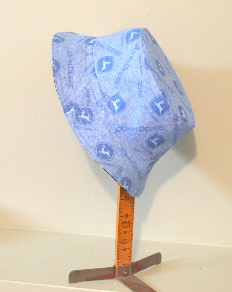May include: A blue bucket hat with a white John Deere logo pattern. The hat is on a wooden stand with a ruler.