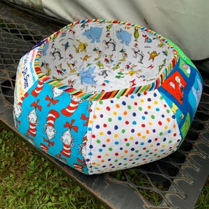 Puede incluir: Un puf de tela colorido y multi-patrón con un fondo blanco y una impresión de Dr. Seuss "El Gato en el Sombrero" en el lateral. El puf está relleno de frijoles y es perfecto para descansar o sentarse.