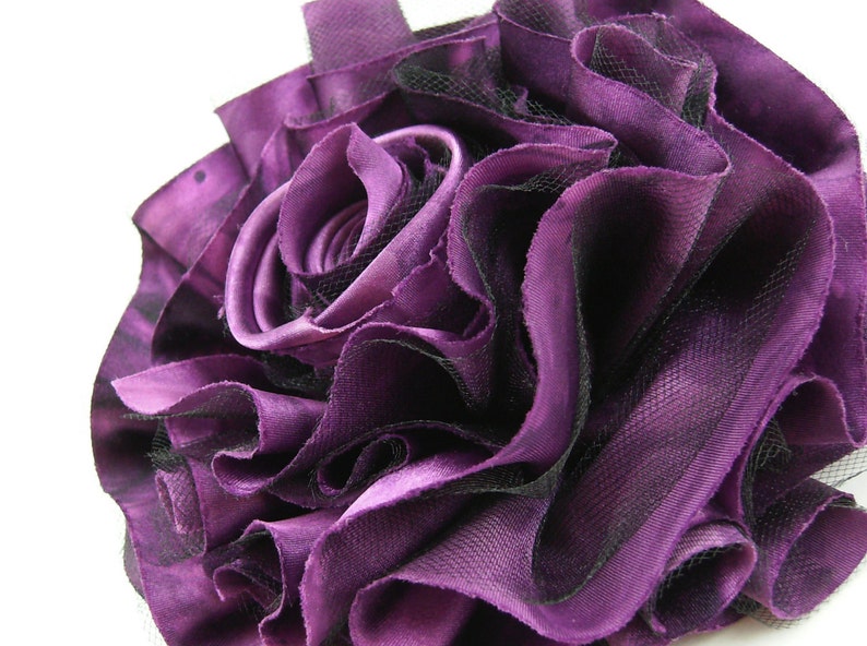 K&ouml;nnte beinhalten: Eine gro&szlig;e, violette Stoffrose mit schwarzem Netz. Die Rose besteht aus mehreren Stofflagen und ist sehr voll.