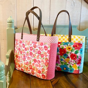 Puede incluir: Dos bolsos tote acolchados con estampados florales y asas de cuero marrón. El bolso de la izquierda tiene un estampado floral rosa y blanco con un acento de lunares rosa. El bolso de la derecha tiene un estampado de cuadros vichy amarillo y blanco con un estampado floral azul y blanco.