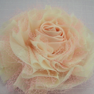 K&ouml;nnte beinhalten: Eine gro&szlig;e, blassrosa und cremefarbene Stoffblume mit einem ger&uuml;schten, geschichteten Design. Die Blume besteht aus einem weichen, durchsichtigen Stoff mit einer zarten Spitzen&uuml;berlagerung.