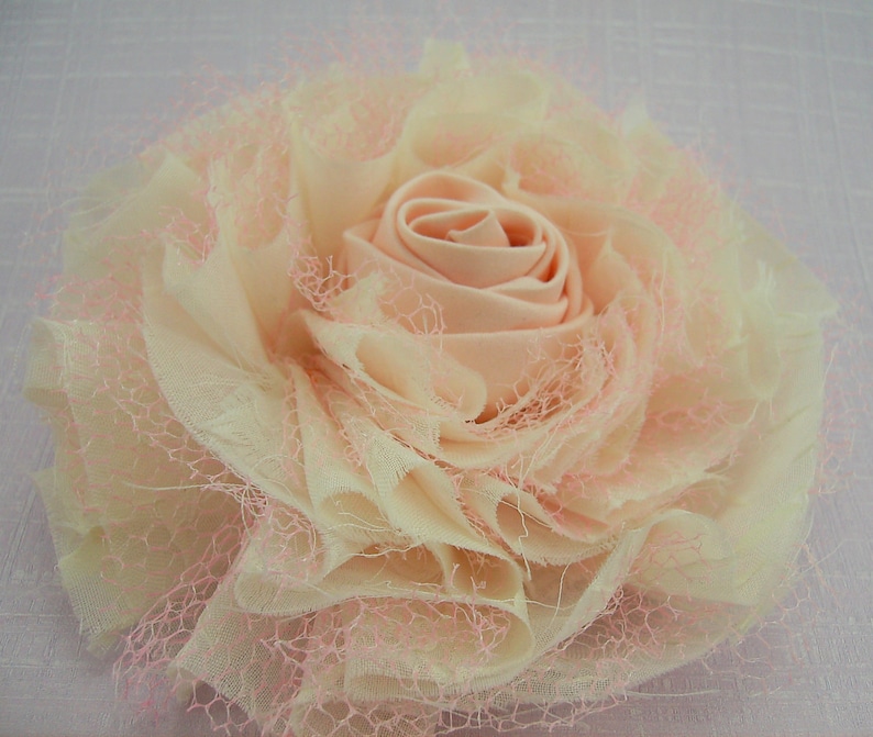 K&ouml;nnte beinhalten: Eine gro&szlig;e, blassrosa und cremefarbene Stoffblume mit einem ger&uuml;schten, geschichteten Design. Die Blume besteht aus einem weichen, durchsichtigen Stoff mit einer zarten Spitzen&uuml;berlagerung.