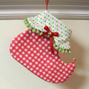 Puede incluir: Un calcetín navideño hecho a mano con forma de zapato de elfo. El calcetín presenta un patrón de cuadros rojos y blancos en el zapato y un puño blanco con lunares verdes. Una cinta roja y una campana añaden detalles festivos.