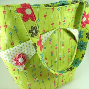 Puede incluir: Una bolsa de tela verde con estampado floral y lunares blancos. La bolsa tiene dos asas y un bolsillo grande en la parte delantera.