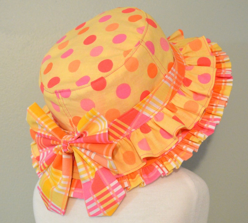 May include: A yellow sun hat with pink and orange polka dots and a pink and orange plaid bow. The hat has a ruffled brim.