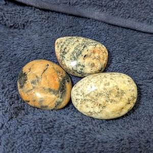 May include: Three smooth, oval-shaped stones with a speckled pattern of black, white, and orange. The stones are arranged in a triangular shape on a blue fabric background.
