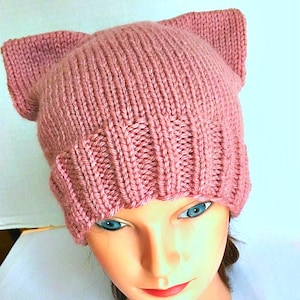 May include: A knitted pink hat with cat ears. The hat is made of a thick yarn and has a ribbed brim. The cat ears are upright and add a playful touch to the hat. The hat is displayed on a mannequin head.