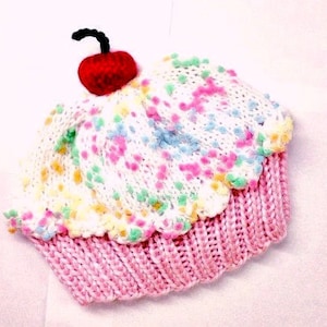 May include: A knitted cupcake hat with a red cherry on top. The hat features a pink base and a white top with colorful sprinkles. The cherry has a black stem. The hat is designed to resemble a cupcake.