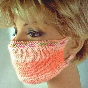May include: A close-up of a knitted face mask in a peach color with a pink and green trim. The mask is being worn by a person with blonde hair.