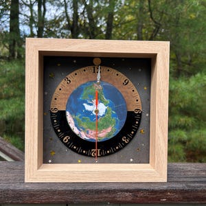 May include: Square wooden clock featuring a detailed Earth globe design at its center. The clock face has a black and brown ring with numbers and star accents. The hands are red and the frame is light wood.