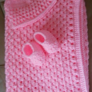 May include: A handmade, light pink crochet baby set, including a blanket, hat, and booties. The blanket and hat have a textured pattern, and the booties have pink soles. The set is designed for a newborn.