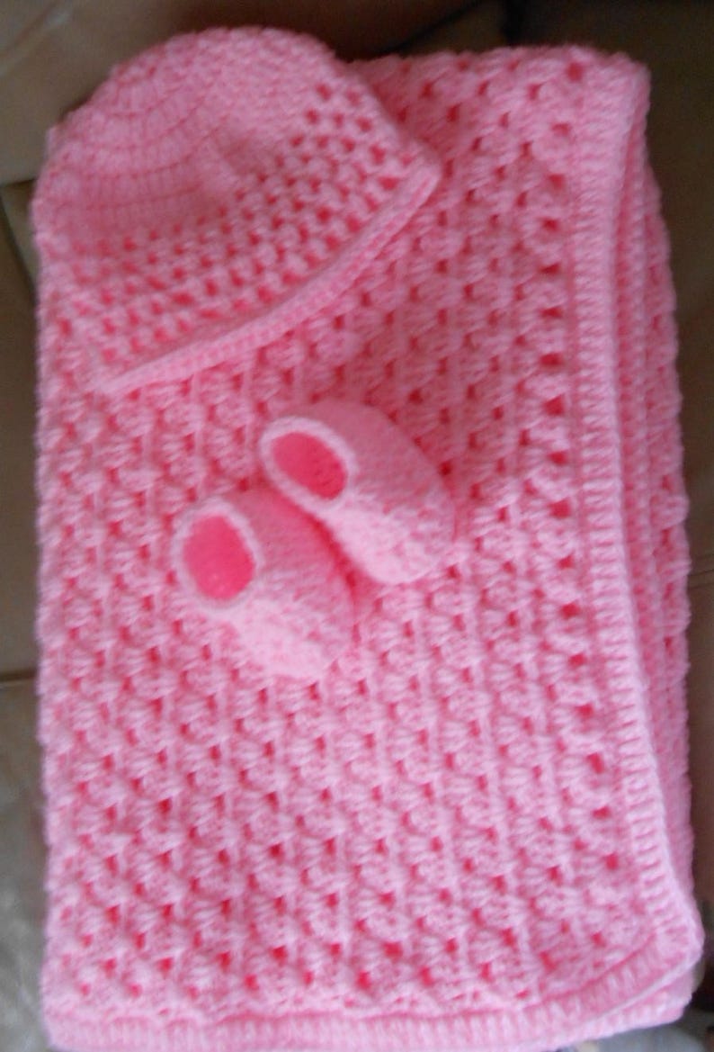 May include: A handmade, light pink crochet baby set, including a blanket, hat, and booties. The blanket and hat have a textured pattern, and the booties have pink soles. The set is designed for a newborn.