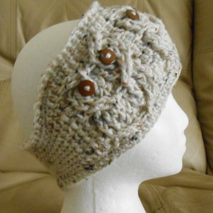 May include: A cream-coloured, hand-crocheted headband with a cable knit design. The headband features three brown wooden buttons and flecks of brown and black throughout the yarn. This cosy accessory is designed to keep the ears warm.