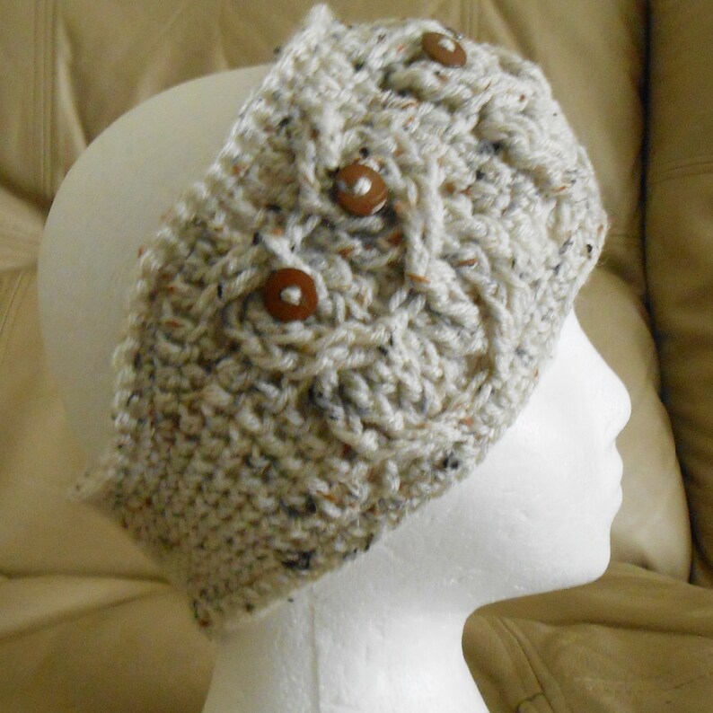 May include: A cream-coloured, hand-crocheted headband with a cable knit design. The headband features three brown wooden buttons and flecks of brown and black throughout the yarn. This cosy accessory is designed to keep the ears warm.
