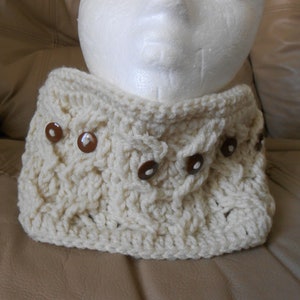 May include: Cream-coloured crocheted cowl with a textured cable knit design. The cowl is adorned with six brown, circular buttons. The neck warmer is displayed on a white mannequin head.