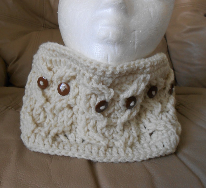May include: Cream-coloured crocheted cowl with a textured cable knit design. The cowl is adorned with six brown, circular buttons. The neck warmer is displayed on a white mannequin head.