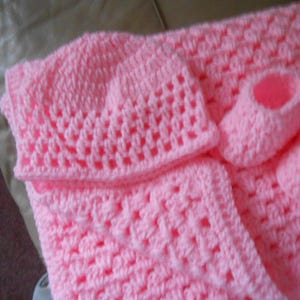 May include: A baby set in a soft pink hue, featuring a crocheted hat, blanket, and two booties. The set is made with a textured stitch, perfect for a newborn. The blanket and hat are square shaped. The booties are small and delicate.