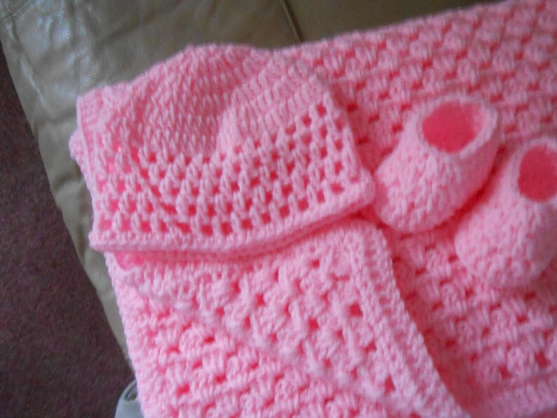 May include: A baby set in a soft pink hue, featuring a crocheted hat, blanket, and two booties. The set is made with a textured stitch, perfect for a newborn. The blanket and hat are square shaped. The booties are small and delicate.