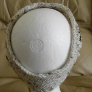 May include: A knitted headband in a neutral colour scheme. The headband is made from a textured yarn with flecks of blue, brown, and black. The headband is displayed on a white mannequin head.