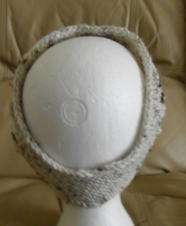 May include: A knitted headband in a neutral colour scheme. The headband is made from a textured yarn with flecks of blue, brown, and black. The headband is displayed on a white mannequin head.