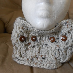 May include: A cream-coloured, hand-knitted cowl with brown buttons. The cowl is made of a textured yarn with flecks of colour. The buttons are round and sewn on the front. This neck warmer is a cosy accessory.