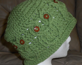 May include: A green crocheted hat with a textured pattern and brown buttons. The hat is displayed on a white mannequin head. The hat is a stylish accessory for cold weather.