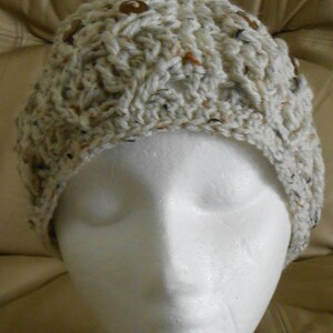 May include: A hand-crocheted hat in off-white with brown buttons. The hat features a textured, speckled pattern with brown, black, and orange flecks. It is displayed on a white mannequin head, highlighting its shape and design. A warm and stylish hat for the colder months.