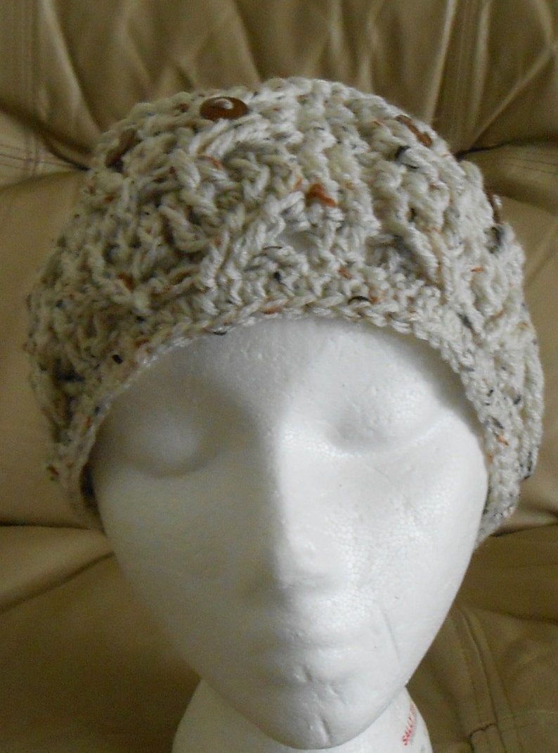 May include: A hand-crocheted hat in off-white with brown buttons. The hat features a textured, speckled pattern with brown, black, and orange flecks. It is displayed on a white mannequin head, highlighting its shape and design. A warm and stylish hat for the colder months.