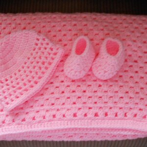 May include: A handmade, light pink crochet baby set, including a blanket, hat, and booties. The blanket is rectangular, with a textured open weave pattern. The hat is round, and the booties are small and delicate. The set is soft and cozy.