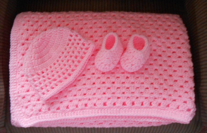 May include: A handmade, light pink crochet baby set, including a blanket, hat, and booties. The blanket is rectangular, with a textured open weave pattern. The hat is round, and the booties are small and delicate. The set is soft and cozy.