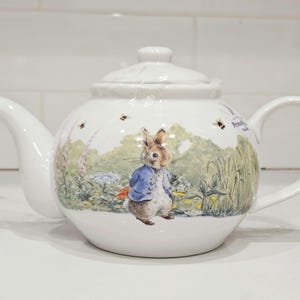 May include: White ceramic teapot with a lid, spout, and handle. The teapot features a colourful illustration of a rabbit in a blue jacket, surrounded by a garden scene with bees. The teapot is designed with a classic, rounded shape.
