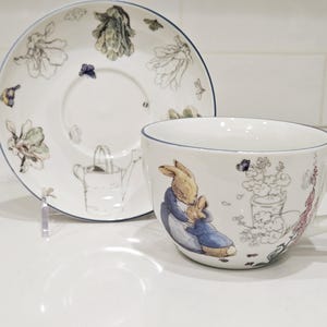 Può includere: Servizio da tè in ceramica bianca con bordo blu. La tazza e il piattino presentano illustrazioni di un coniglio, fiori e un annaffiatoio. Il design ha un aspetto stravagante e fiabesco.