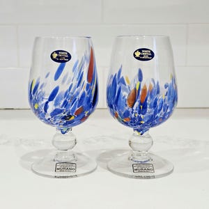 May include: Two clear wine glasses with blue, red, and yellow abstract designs. Each glass has a round base, a stem with a bulbous detail, and a rounded bowl. The glasses have labels that read "WHITE CRISTAL" and "MURANO".