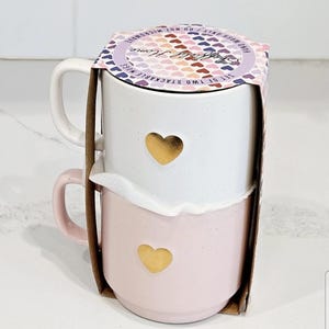 Pode incluir: Um conjunto de duas canecas empilháveis, uma branca e outra rosa, com detalhes de coração dourado. A caneca branca é empilhada em cima da caneca rosa. As canecas são embaladas em um suporte de papelão com o texto "Simply Home".